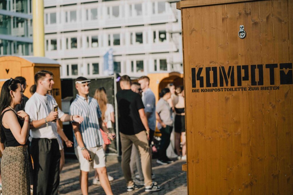 Festivalbesucher vor Holztoiletten – Menschen laufen über ein belebtes Festivalgelände mit Toilettenkabinen aus Holz im Hintergrund. Auf einer nahen Kabine auf der rechten Bildseite kann man "Kompotoi - Die Komposttoilette zum Mieten) lesen.