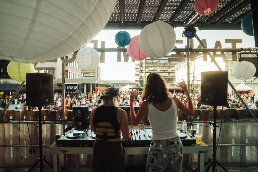 DJ-Duo auf einer Open-Air-Bühne – Zwei weibliche DJs stehen hinter einem Mischpult und legen auf, während Papierlaternen über ihnen hängen. Man sieht von beiden den Rücken und dahinter die feinernde Menge.