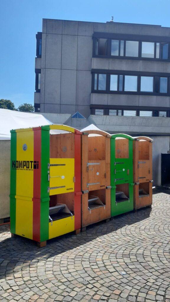 Toilettenstation auf dem Festivalgelände – Mehrere Toilettenkabinen aus Holz stehen nebeneinander auf einem gepflasterten Platz mit einem Gebäude im Hintergrund. Zwei Kabinen sind holzfarben, zwei bunt angemalt in Rasta-Farben.